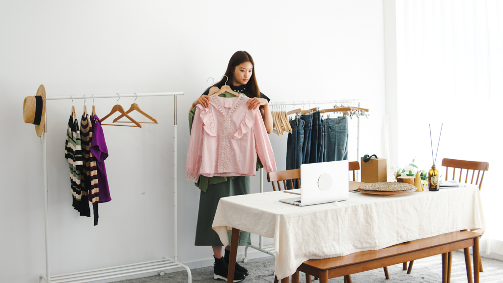 A woman stands in a room holding up two pink blouses, surrounded by clothing racks, a laptop on a table, and various items—capturing the essence of a closet cleanout guide in action.